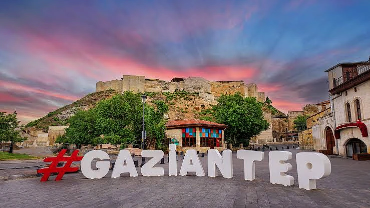 Gaziantep kalesi arka planda, ön planda büyük harflerle #GAZIANTEP yazısı ve yeşil ağaçlar.