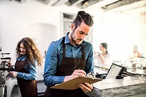 Kafe ortamında çalışan bir adam, not alırken. Arka planda başka çalışanlar var.
